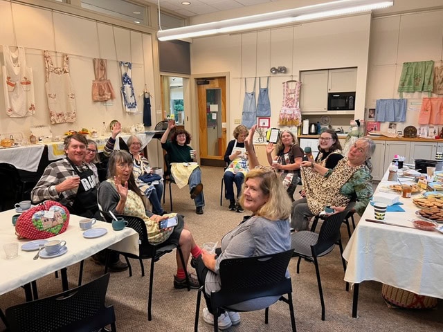 a group of people sit around tables with food and drinks in a room decorated with aprons, smiling and gesturing toward the camera.