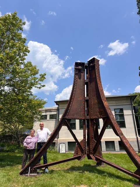 two people stand next to a large, rusted metal structure outdoors, in front of a modern building and a tree under a partly cloudy sky.