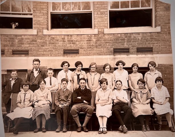 a black and white photo of a group of young women and three men posing in two rows outside a brick building with large windows.