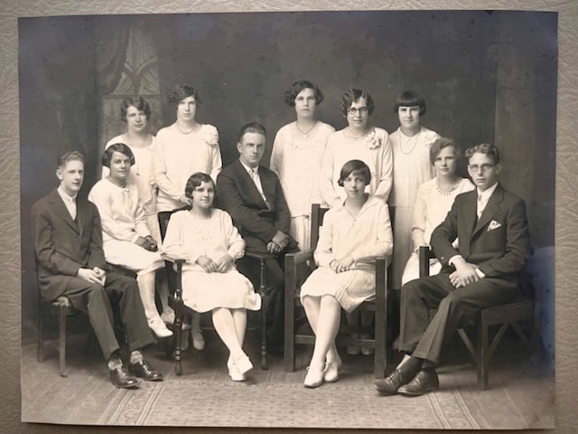 a black and white portrait of nine women and three men posing in formal attire, arranged in two rows, against a plain studio backdrop.