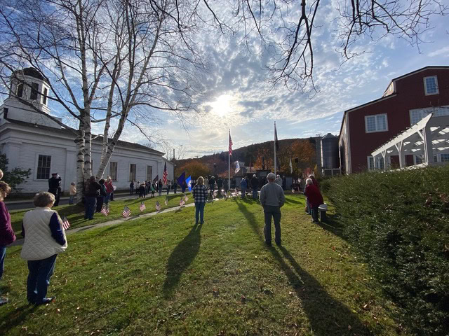 people gather outdoors near a white building and flagpoles on a sunny day, with american flags visible on the lawn and fall trees in the background.