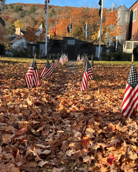 rows of small american flags are placed in fallen autumn leaves leading up to a memorial area with multiple flagpoles and a red building in the background.