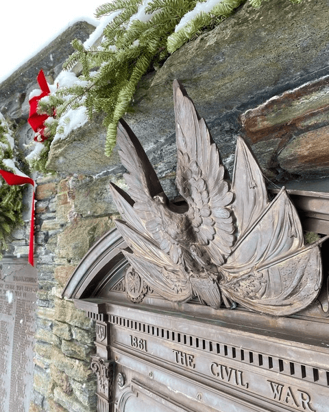 bronze eagle sculpture with spread wings mounted on a stone wall, decorated with evergreen branches and red ribbons, with snow visible.