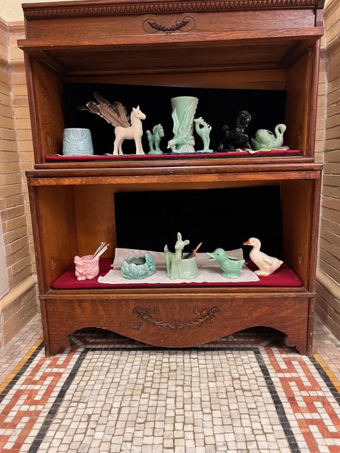 wooden cabinet with two shelves displaying various ceramic animal and plant figurines, including swans, ducks, and a unicorn, all arranged on red and white cloths.