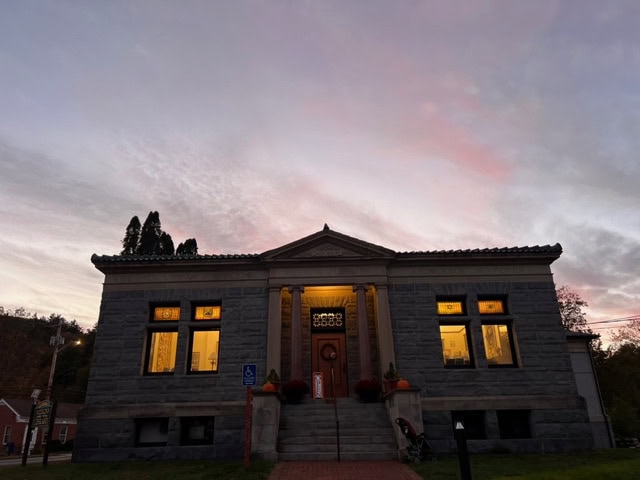 a stone building with large windows is lit from within at dusk, with a pink and purple sky in the background. the entrance has columns and a small set of stairs.