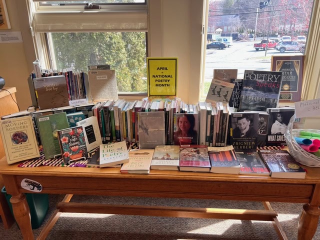 a library display table features poetry books and a sign reading "april national poetry month" in front of windows with a view of trees and a street outside.
