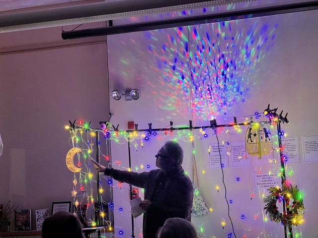 a person stands in front of a wall decorated with colorful string lights, holding a crescent moon shaped light, with multicolored reflections on the wall.