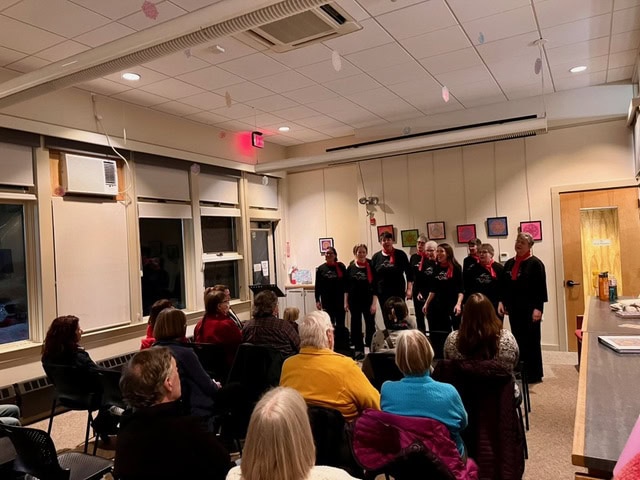 a group of performers sing in front of an audience in a small, well lit room with art on the walls and ceiling decorations.