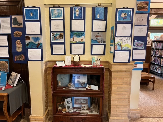 a wooden display case with artifacts stands below a wall exhibit of artwork and poems at a library, with bookshelves visible in the background.