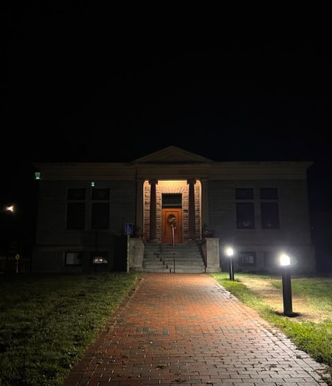 a brick walkway leads to the entrance of a small, columned building at night, illuminated by outdoor lights and surrounded by grass.