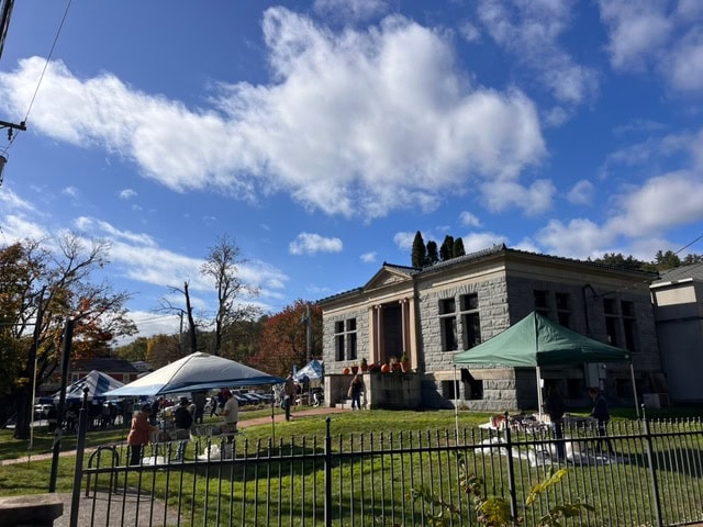 people gather under tents on a lawn in front of a historic stone building with columns on a sunny day with scattered clouds.