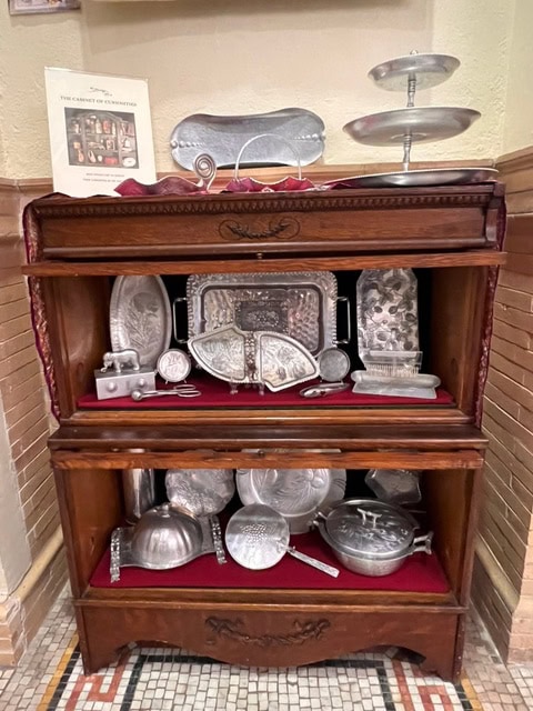 a wooden display cabinet with glass shelves holds an assortment of silver trays, dishes, serving utensils, and decorative metalware, all arranged on red velvet fabric.