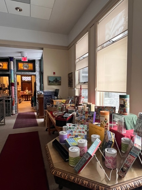 a display table with assorted gift items and stationery is set near large windows inside a well lit hallway of a building, with a doorway visible in the background.
