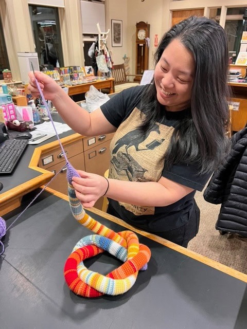 a woman stands at a desk, smiling as she crochets with purple yarn. a colorful, striped knitted object shaped like a snake rests in front of her.