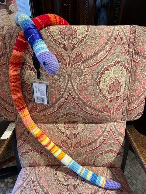 a long, multicolored crocheted snake is draped over a patterned upholstered chair with a badge attached near its head.