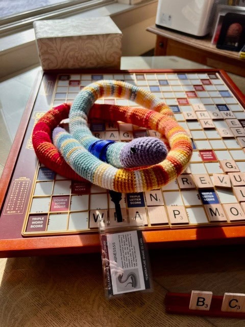 a colorful knitted snake rests on a scrabble board, with letter tiles and a name badge visible nearby on a wooden table.