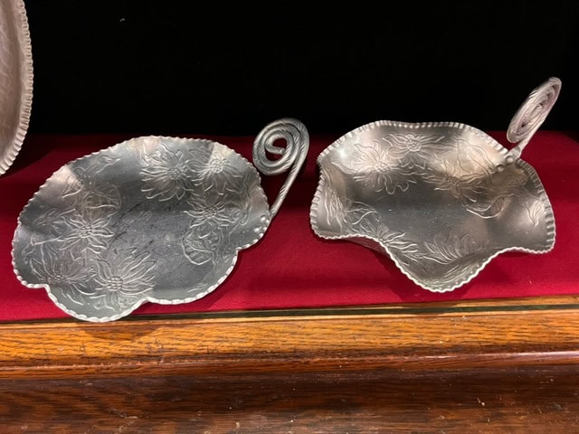 two decorative silver dishes with engraved floral patterns and coiled handles displayed on a red cloth surface.
