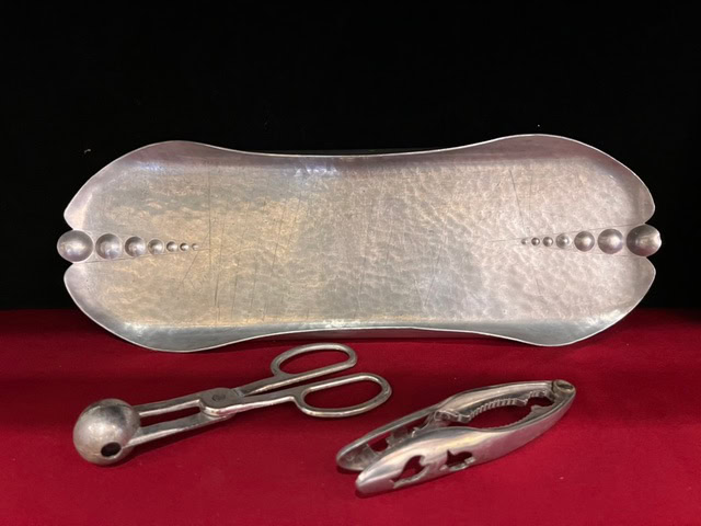 a metal tray with a hammered texture is displayed behind a pair of metal tongs and a nutcracker, all placed on a red surface.