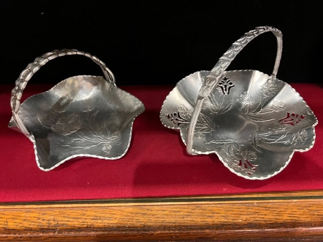 two small, ornate silver baskets with floral designs and handles are displayed on a red fabric surface.