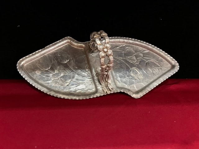 an ornate, silver toned divided dish with floral engravings and a decorative handle, displayed on a red surface against a black background.