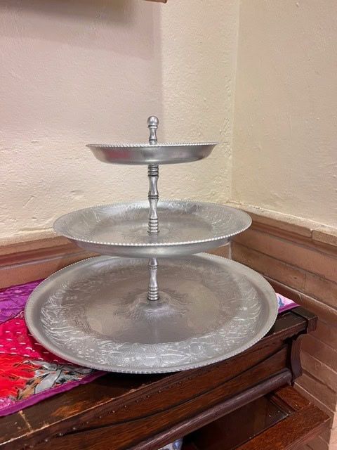 a three tiered silver serving tray is displayed on a wooden surface against a beige wall.