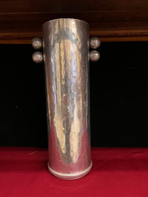 a tall, cylindrical, hammered metal vase with four round knobs, two on each side, placed on a red surface against a dark background.