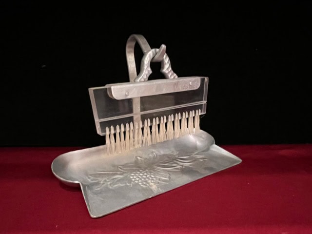 a metal nutcracker with a handle and pick holder sits on a metal tray embossed with a grape design, against a dark background and red surface.