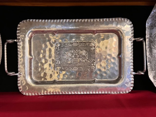 rectangular hammered metal tray with raised handles and a central embossed fruit motif, displayed on a red surface against a black background.