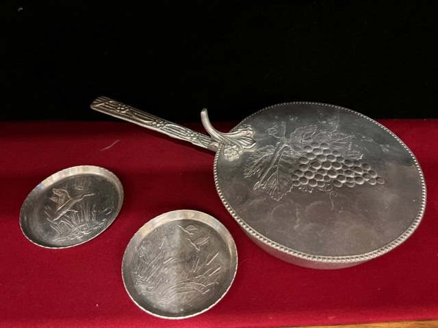 a silver hand mirror with a grapevine design and two round silver dishes with nature engravings are displayed on a red surface.