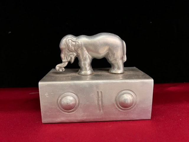 a metallic elephant figurine stands atop a rectangular metal block with two round protrusions and a vertical line, set against a red and black background.