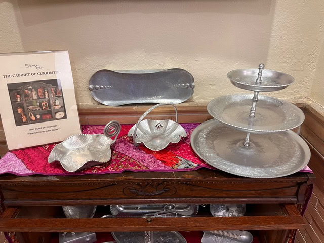 handcrafted silver serving dishes and trays displayed on a wooden table, with a framed sign titled "the cabinet of curiosities" on the left side.