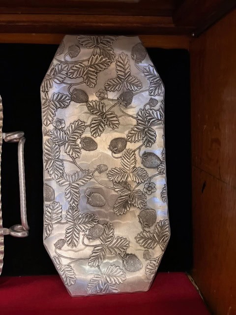 rectangular silver colored tray with rounded corners, decorated with an embossed pattern of strawberries, leaves, and stems. the tray is placed inside a wooden display case.