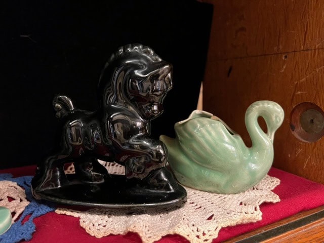 a black ceramic horse figurine and a green ceramic swan sit on a lace doily atop a red surface, against a dark background.