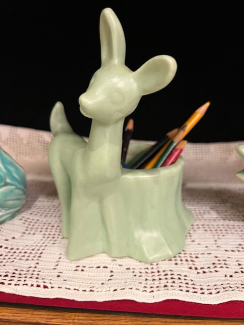 a pale green ceramic deer shaped holder contains colored pencils, placed on a white lace doily with a black background.