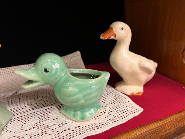 two ceramic duck figurines, one green with an open beak and the other white with an orange beak and feet, are displayed on a lace doily atop a red cloth.