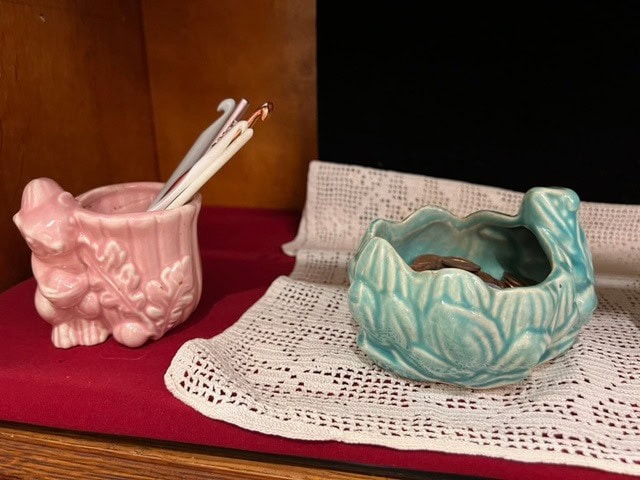 a pink ceramic container holding crochet hooks sits next to a blue ceramic bowl with coins inside, both on a crocheted doily atop a wooden surface.