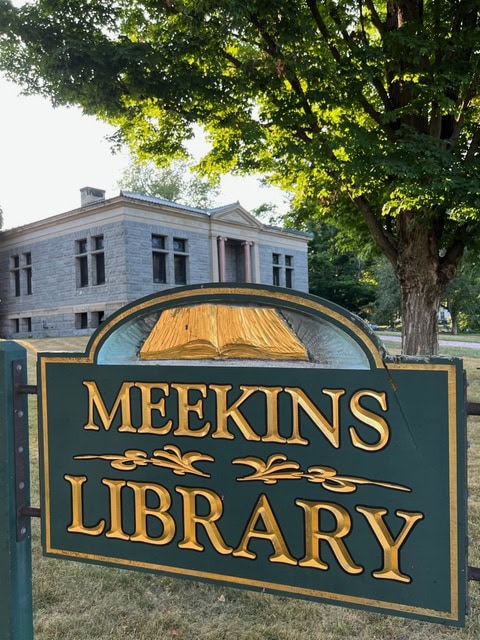 a green and gold sign reading "meekins library" stands in front of a stone library building with columns and large windows, surrounded by trees.