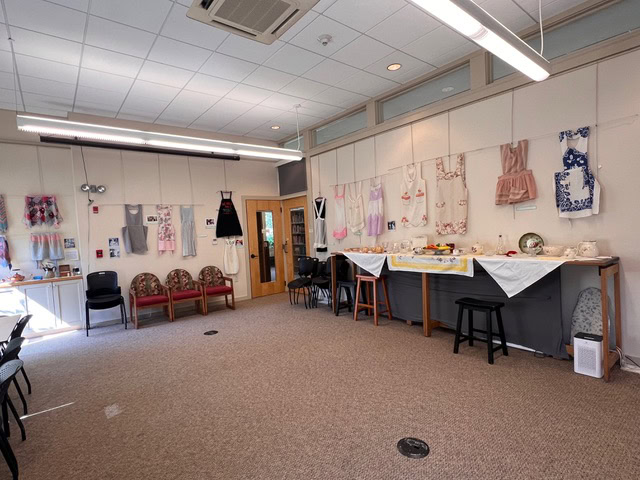 a room with aprons hanging on the walls, tables set with dishes and linens, chairs along the sides, and fluorescent lighting overhead.