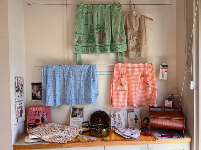 a display of vintage aprons, sewing patterns, books, and various sewing accessories arranged on shelves and hanging on a wall.