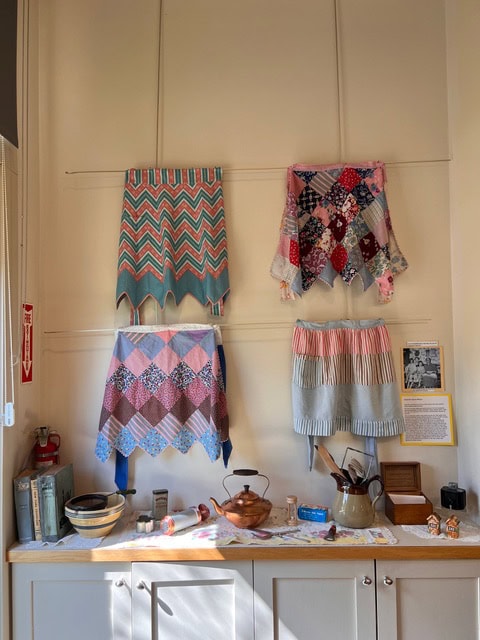 four vintage aprons are displayed on a cream wall above a counter with bowls, utensils, a teapot, and informational placards, all in natural sunlight.