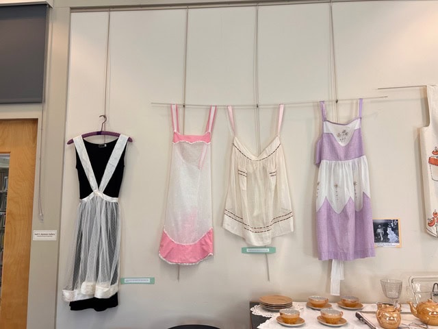 four vintage aprons in black, white, pink, and lavender hang on a wall above a table set with plates and glassware.