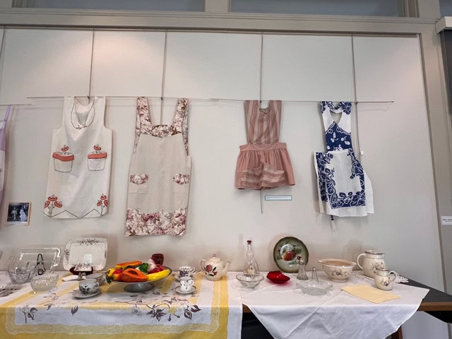 three vintage aprons hang on a wall above a table set with assorted dishes, teacups, glassware, and decorative ceramics.