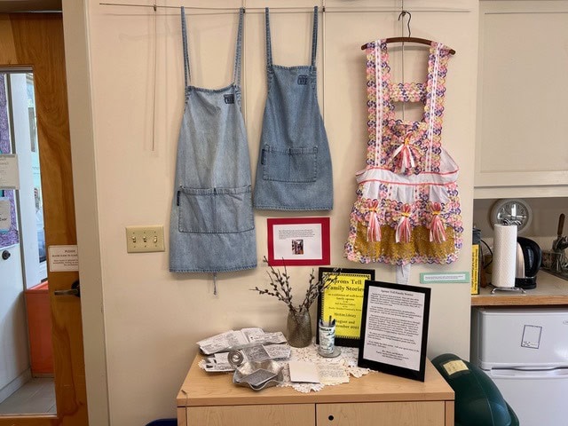three aprons hang on a wall above a display table with books, a vase, and informational signs in a kitchen or classroom setting.