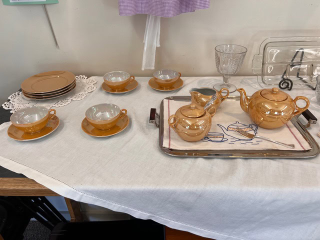 a set of amber colored tea cups with saucers, a teapot, creamer, and sugar bowl arranged on a tray with a white embroidered cloth, next to stacked plates and a glass.