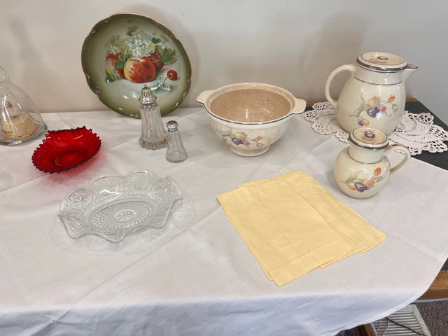 a table with a white cloth displays vintage ceramic pitchers, a mixing bowl, glass dishes, salt and pepper shakers, a red glass bowl, a plate with fruit design, and a folded yellow napkin.
