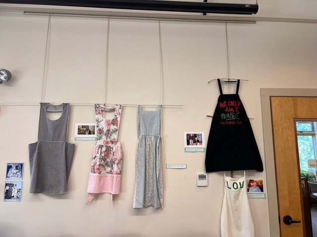 four aprons hang on a wall above photos and captions; one apron is black with red and green writing, while the others are patterned in light colors and florals.