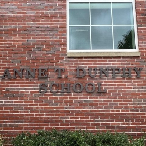 brick exterior wall of a building with large letters spelling "anne t. dunphy school" below a window. shrubs are visible at the bottom of the image.