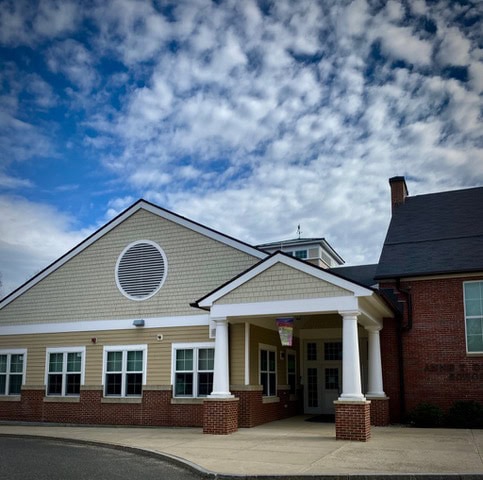a one story building with tan siding, white trim, and brick accents, featuring a columned entrance under a partly cloudy sky.