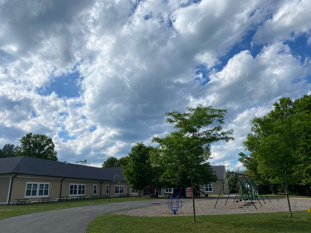 a playground with swings and climbing equipment next to a building, with a path and trees under a partly cloudy sky.