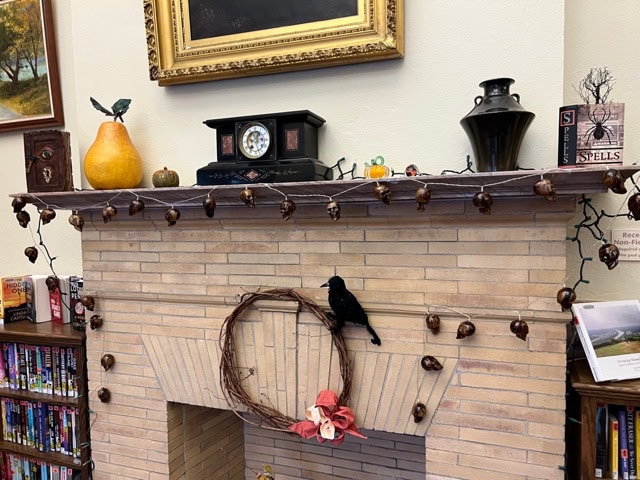a mantel decorated with halloween themed items, including a faux crow, mini pumpkins, skull string lights, a pear, books, and a clock, above a brick fireplace.